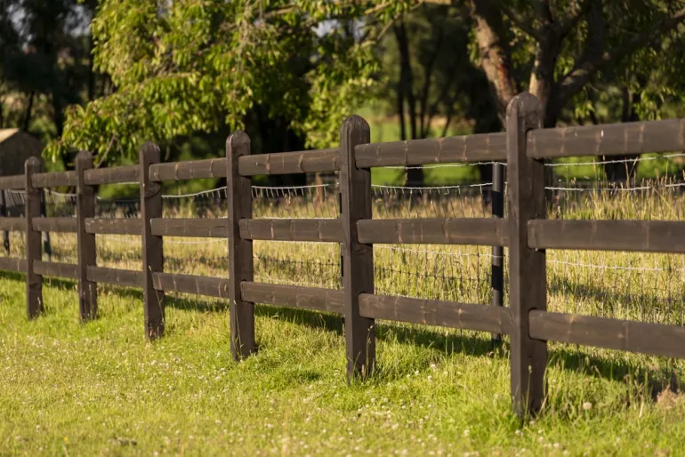 Une clôture en bois noir avec des poteaux carrés et 3 lisses entourant un champ