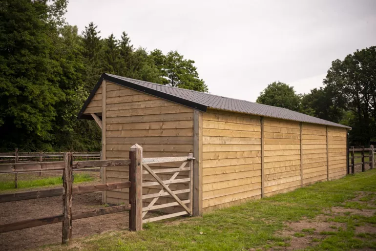The back of four wooden horse shelters with black roof tiles on a field with wooden fences with three rails